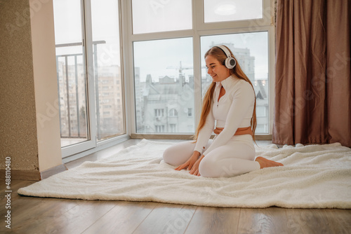 Side view portrait of relaxed woman listening to music with headphones lying on carpet at home. She is dressed in a white tracksuit.