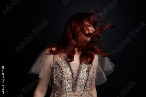portrait of pretty female model with red hair wearing glamorous fantasy tulle gown and crown.  Posing with a moody dark background. © faestock