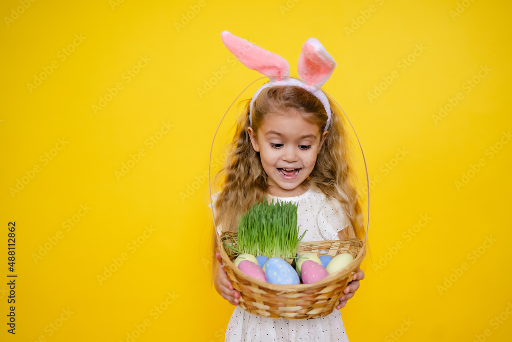 A cheerful girl with rabbit ears on her head with a basket of colored eggs in her hands on a yelow background. Funny crazy happy baby. Easter to the child. A cheerful girl with rabbit ears on her head with a basket of colored eggs in her hands on a yelow background. Funny crazy happy baby. Easter to the child.