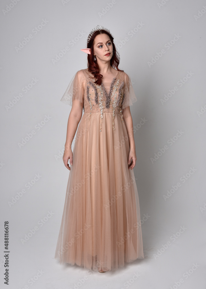  Full length portrait of pretty female model with red hair wearing glamorous fantasy tulle gown and crown.  Posing with gestural arms on a studio background
