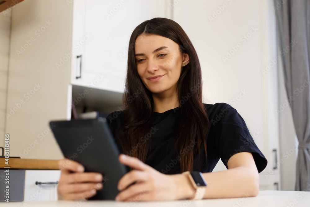 Cheerful white woman browsing internet on laptop computer in home kitchen. Happy brunette female with long dark hair using modern device for online communication and entertainment during lockdown Cheerful white woman browsing internet on laptop computer in home kitchen. Happy brunette female with long dark hair using modern device for online communication and entertainment during lockdown