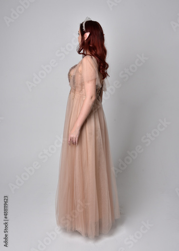 Full length portrait of pretty female model with red hair wearing glamorous fantasy tulle gown and crown. Posing with gestural arms on a studio background