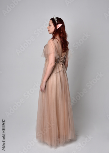 Full length portrait of pretty female model with red hair wearing glamorous fantasy tulle gown and crown. Posing with gestural arms on a studio background