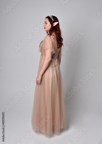 Full length portrait of pretty female model with red hair wearing glamorous fantasy tulle gown and crown. Posing with gestural arms on a studio background