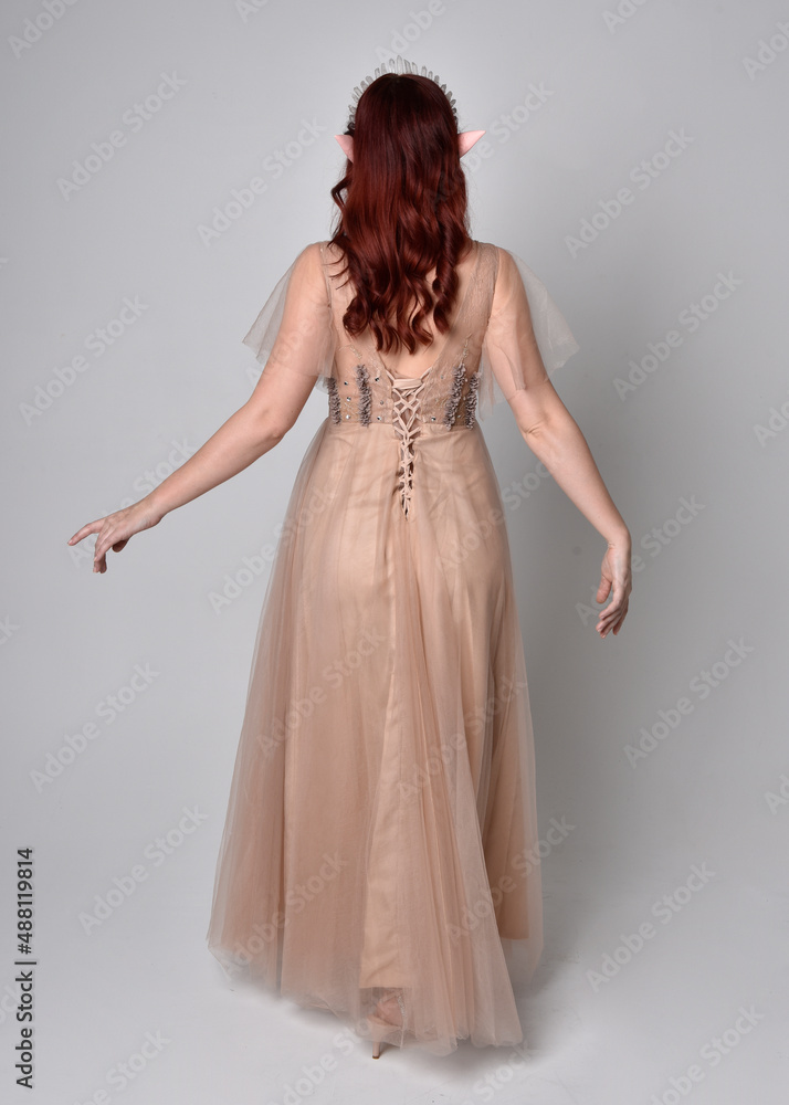 Full length portrait of pretty female model with red hair wearing glamorous fantasy tulle gown and crown.  Posing with gestural arms on a studio background