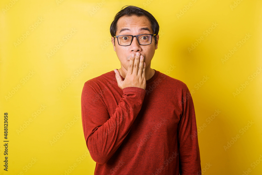 Casual man in glasses covering mouth with hands, surprised expression isolated on yellow background Casual man in glasses covering mouth with hands, surprised expression isolated on yellow background