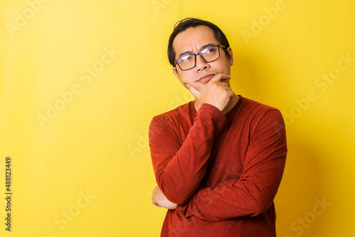 Casual man in glasses with imaginative expression with hand holding chin isolated on yellow background © Gatot