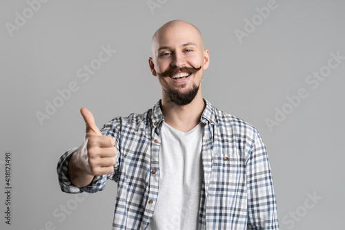 Smiling bald bearded with mustache man in checkered shirt gestures thumb up over gray background