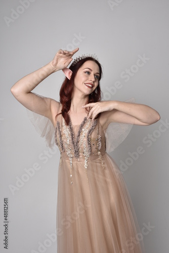 portrait of pretty female model with red hair wearing glamorous fantasy tulle gown and crown. Posing with gestural arms on a studio background