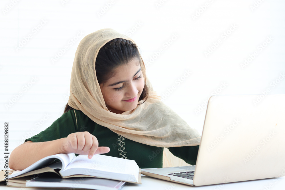 Adorable smiling Pakistani Muslim girl with beautiful eyes wearing hijab, studying online, doing homework by using laptop computer, student kid reading book on white background. Adorable smiling Pakistani Muslim girl with beautiful eyes wearing hijab, studying online, doing homework by using laptop computer, student kid reading book on white background.