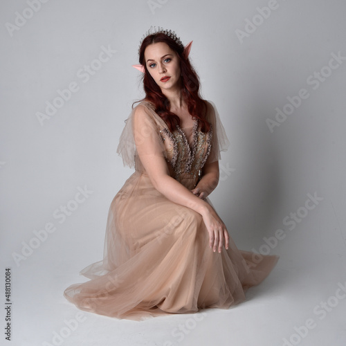 Full length portrait of pretty female model with red hair wearing glamorous fantasy tulle gown, crown and shroud veil. Posing in a seated kneeling pose on a studio background