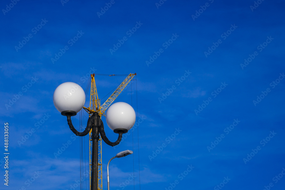 A yellow construction crane against a blue sky behind a black metal pole and white street lights