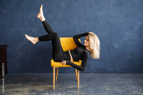 An elegant blonde girl lies impressively on a chair. Yoga style. Unusual poses for portraits. Good stretching and grace