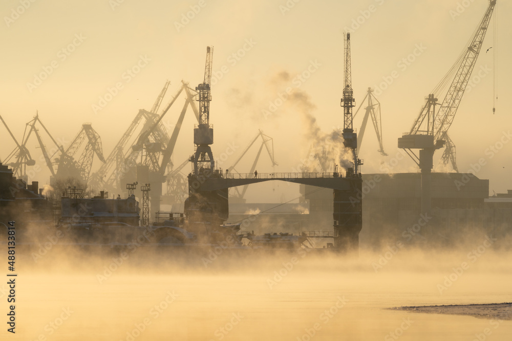 Obraz premium Cranes of Baltic shipyard in St. Petersburg in frosty winter day, steam over the Neva river, smooth surface of the river at sunset.