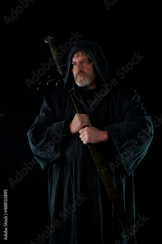 Portrait of courageous monk-warrior in black cloak with sword in hands on black background. Historical medieval templier fantasy