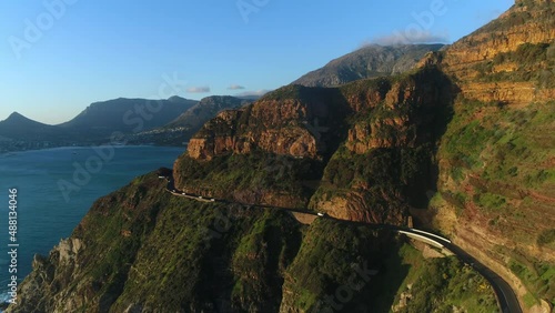 Aerial view of cars driving along Chapman's peak mountain pass while the sun is setting. Camera tracks forward to he road and slowly ascends over one of the peaks.