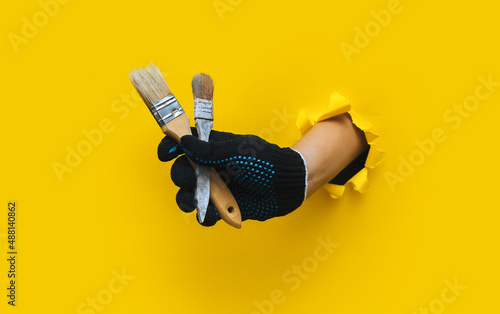 The right painter's hand in a black cloth knitted glove holds a two bristle brush. Torn hole in yellow paper. The concept of a worker, a labor migrant, a master of his craft. Copy space.