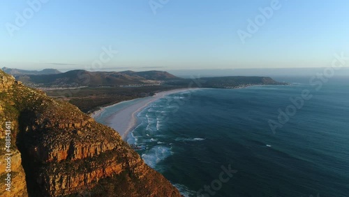Aerial view of Noordhoek's large white beach and flying back to reveal the scenic mountain pass of Chapman's peak. People taking a sunset drive along one of the most iconic coastal roads in the world.
