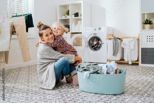 Mother sits on laundry room floor with little daughter, a cute little girl with blonde hair tied up in a bun grabs the woman by neck hugs her tightly they spend time doing housework sorting clothes.