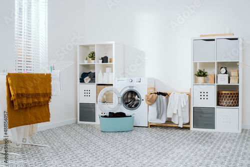 View of home laundry room, dresser with softeners, powder, towels, open washing machine with empty drum, next to wicker basket with colorful laundry items, dryer photo