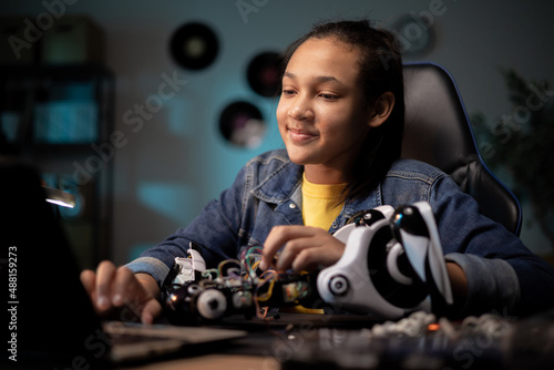 A teenager sits at desk in room in the evening, scrolling through videos, instructions on laptop, browsing the internet for tips on repairing her robot © ABCreative