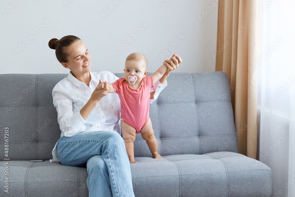 Indoor shot of happy satisfied woman with bun hairstyle wearing white shirt and jeans playing with her toddler daughter in pink bodysuit, maternity leave. Indoor shot of happy satisfied woman with bun hairstyle wearing white shirt and jeans playing with her toddler daughter in pink bodysuit, maternity leave.