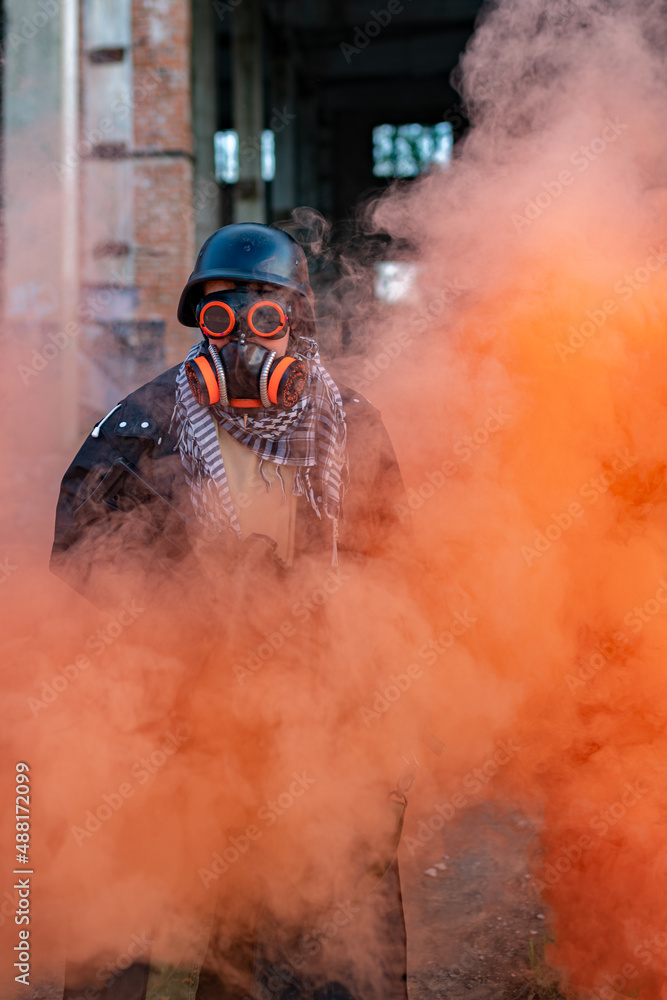 Obraz premium A soldier after a nuclear apocalypse among the ruins: wearing a helmet, steampunk glasses, a gas mask, and a raincoat. In his hands he holds a machine gun, a red smoke screen around him
