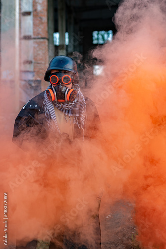 A soldier after a nuclear apocalypse among the ruins: wearing a helmet, steampunk glasses, a gas mask, and a raincoat. In his hands he holds a machine gun, a red smoke screen around him