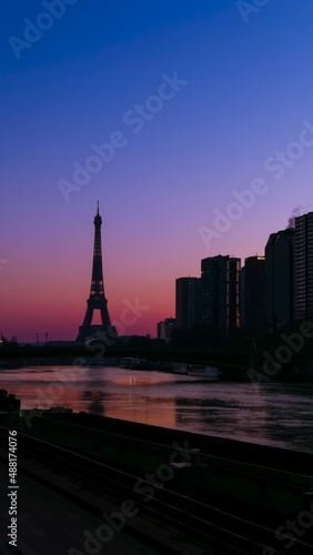 View on the Eiffel tower with group of modern buildings in front of the water of Seine river. Dramatic sky with colorful clouds. Silhouette of a cityscape at sunset or night. © Bruno