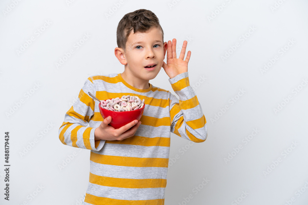 Naklejka premium Little caucasian boy holding a cereal bowl isolated on white background listening to something by putting hand on the ear
