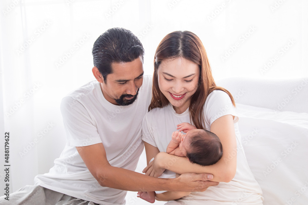 Fototapeta premium Selective focus of Asian man and woman holding newborn baby in bed at home. Happy family smiling with adorable infant, young parents holding little sweet toddler baby, mom hold toddler with love.