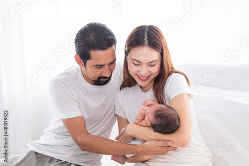 Selective focus of Asian man and woman holding newborn baby in bed at home. Happy family smiling with adorable infant, young parents holding little sweet toddler baby, mom hold toddler with love. © Rakchanok