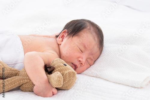 Happy Asian newborn baby sleeping in bed and hug favourite bear doll. Sweet adorable infant sleep in bed in white room with toys comfortable and safety.