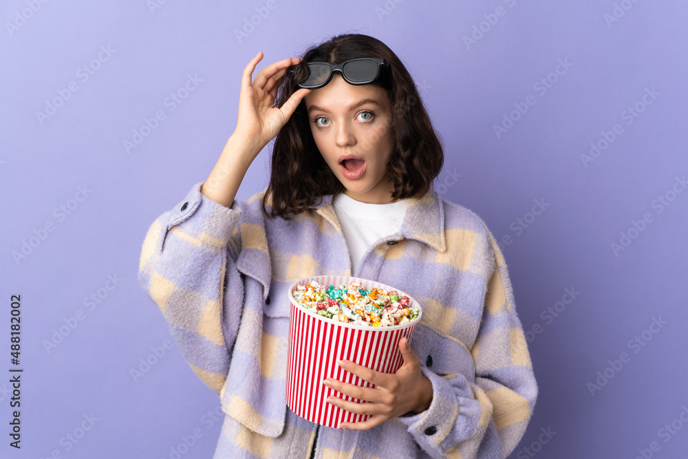 Teenager Ukrainian girl isolated on purple background surprised with 3d glasses and holding a big bucket of popcorns