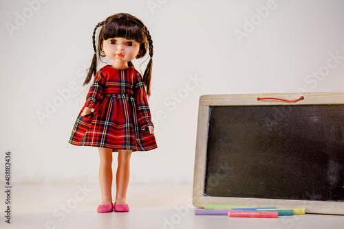 Lovely doll with red dress and pink shoes standing next to the toy wooden blackboard in front of white background