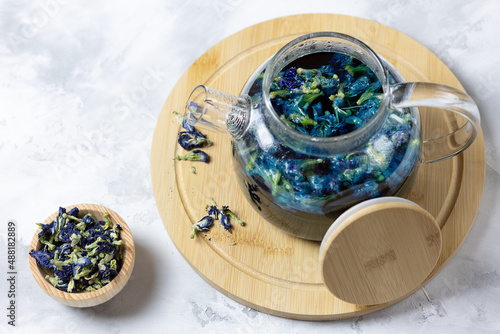 A glass teapot made of organic blue anchana on a light table and a cup with dried tea flowers. Herbal Tea