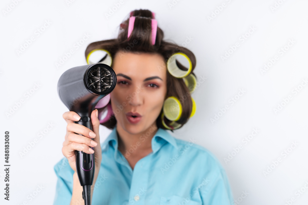 Beautiful housewife. Young cheerful woman with hair curlers and hair dryer getting ready for a date night. Makes a hairstyle at home on a white background. Beautiful housewife. Young cheerful woman with hair curlers and hair dryer getting ready for a date night. Makes a hairstyle at home on a white background.