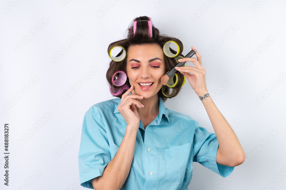 Beautiful housewife. Young cheerful woman with hair curlers, bright pink make-up and a cotouring brush is preparing for a date night, dinner, on a white background. Beautiful housewife. Young cheerful woman with hair curlers, bright pink make-up and a cotouring brush is preparing for a date night, dinner, on a white background.