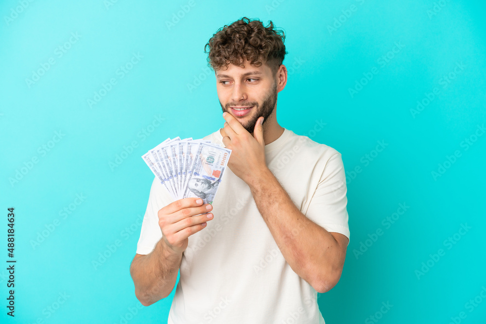 Young caucasian man taking a lot of money isolated on blue background looking to the side and smiling Young caucasian man taking a lot of money isolated on blue background looking to the side and smiling