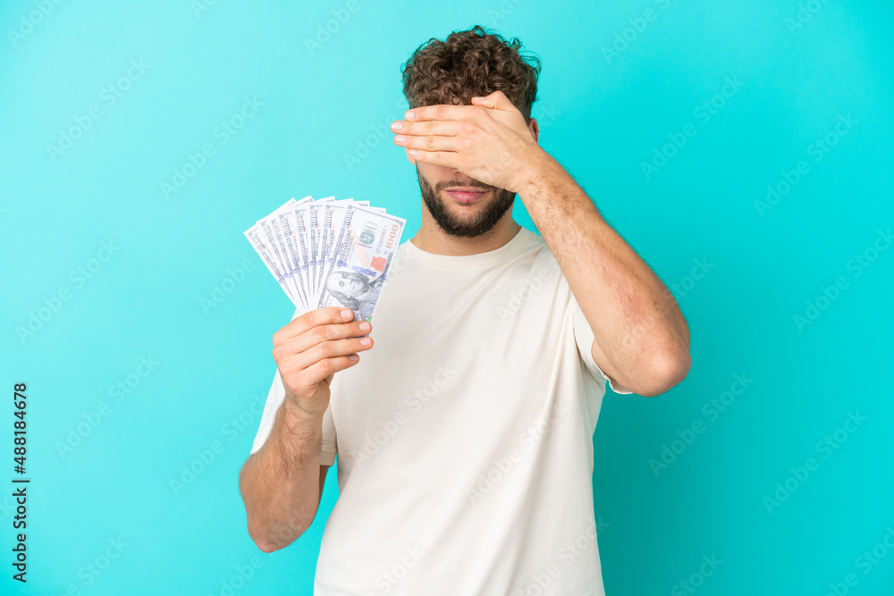 Young caucasian man taking a lot of money isolated on blue background covering eyes by hands. Do not want to see something Young caucasian man taking a lot of money isolated on blue background covering eyes by hands. Do not want to see something