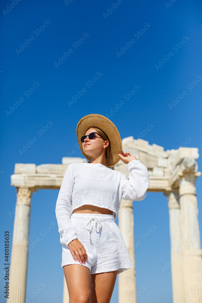 A young slender tanned woman in white shorts and a T-shirt, wearing sunglasses and a hat. Walk on a hot sunny day