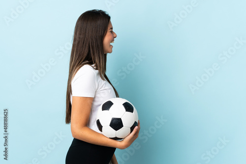 Young football player brazilian girl isolated on blue background laughing in lateral position © luismolinero