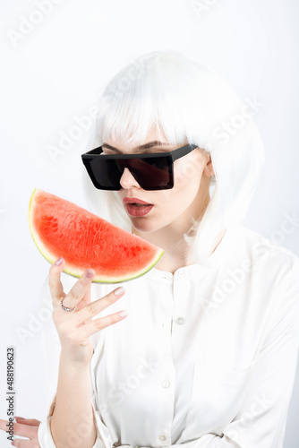 Fashion, abstract and style concept. Close-up studio shot of fancy and beautiful looking woman with white wig and big sunglasses holding slice of red watermelon in her hands. Model with white blouse