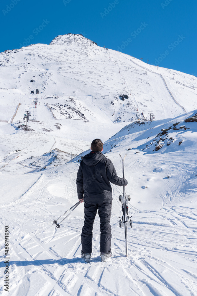 Skier holding his skis in the middle of the snowy mountain looking at the highest peak