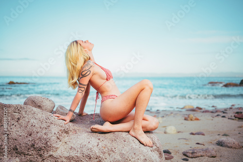 blonde woman in a pink bikini seating on a rock near the beach 