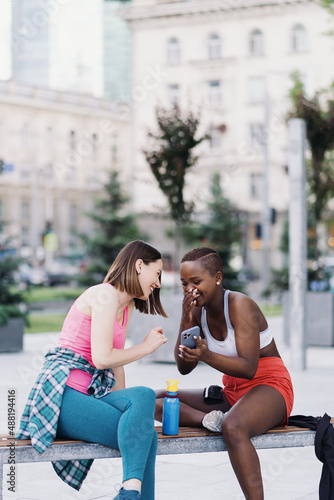 Cheerful smiling friends in sportswear sitting on a bench in the city discussing while using smartphone looking to screen. Multiethnic women having a fitness workout break.
