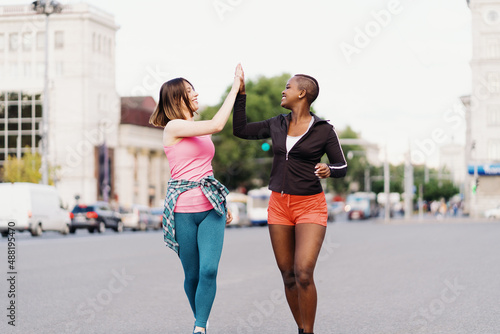 Smiling cheerful friends in sportswear running in the city discussing, giving hi five, celebrating success and happiness. Multiethnic women having a fitness workout.