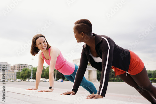 Two smiling diverse young woman in Athletic Workout Clothes are Doing a Plank Exercise working out together in city square.