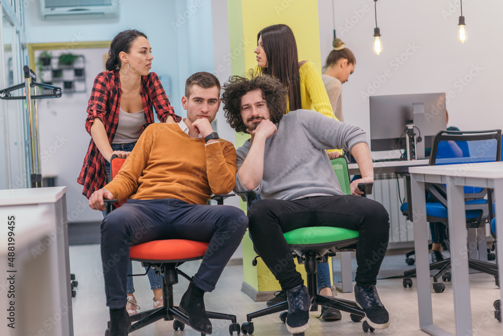 Team building and office fun.Four young cheerful businesspeople in smart casual wear having fun while racing on office chairs and smiling.. Team building and office fun.Four young cheerful businesspeople in smart casual wear having fun while racing on office chairs and smiling..