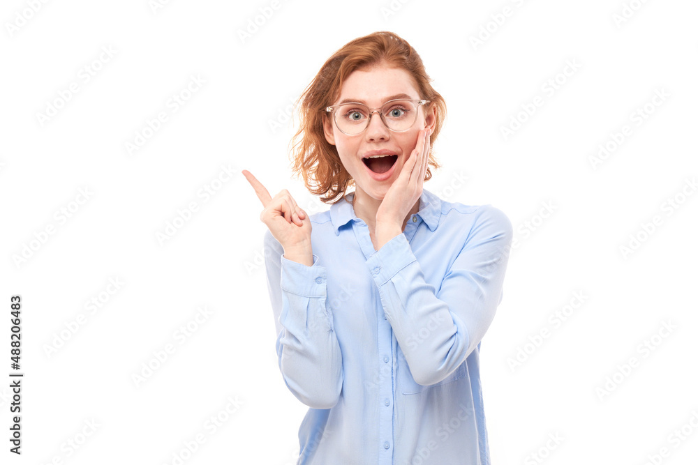 Shocked face red-haired girl in business shirt looks surprised with open mouth on white studio background points finger at copy space Shocked face red-haired girl in business shirt looks surprised with open mouth on white studio background points finger at copy space